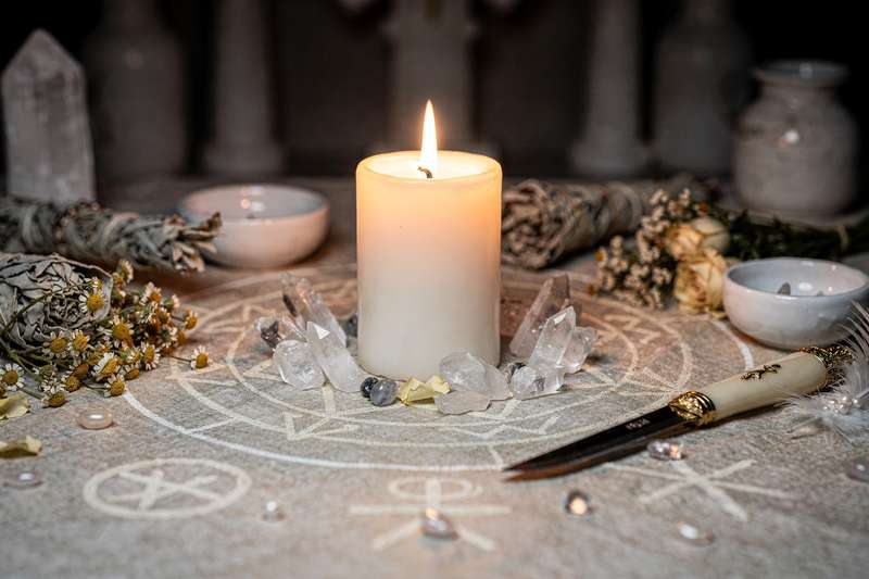 Brennende weiße Kerze auf dunklem Hintergrund, umgeben von Kristallen und Kräutern für spirituelle Rituale und Schutzmagie.